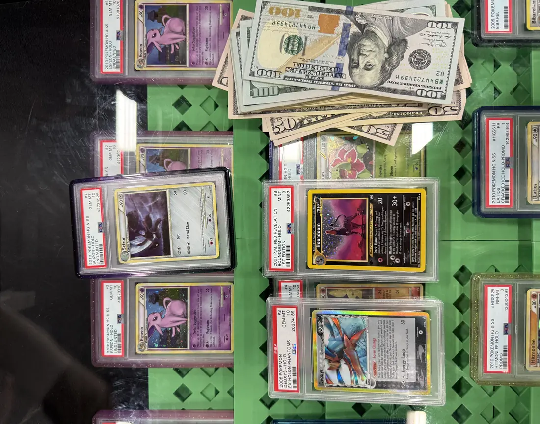 A vendor case at a show: PSA-slabbed Pokémon cards laid out on green foam — Espeon, Scizor, a 1st Edition Houndoom, Deoxys, Latios — with a folded stack of fifty and hundred dollar bills sitting on top of them.